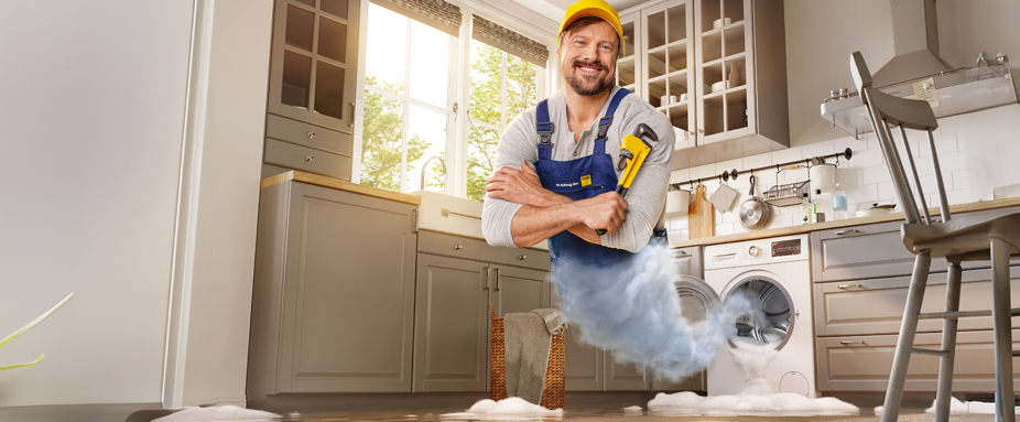 Handwerker mit Rohrzange erscheint aus auslaufender Waschmaschine - ADAC Zuhause-Schutzbrief